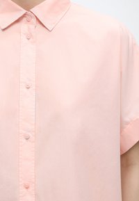 Ανοιχτό ροζ πουκάμισο με κοντά μανίκια και κουμπιά, με μυτερό γιακά, εμφανιζόμενο από το στήθος μέχρι τον ώμο σε ένα άτομο μπροστά από απλό φόντο.