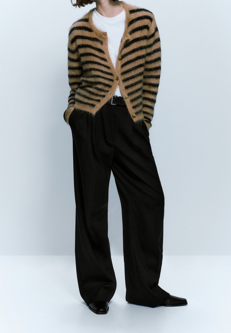 Cardigan rayé beige et noir avec un col rond et fermeture à boutons, associé à un pantalon noir à jambes larges et des chaussures noires pointues.