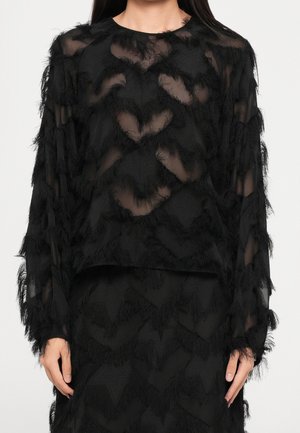 Femme portant un haut noir transparent à manches longues avec des franges en zigzag texturées et une jupe noire assortie.