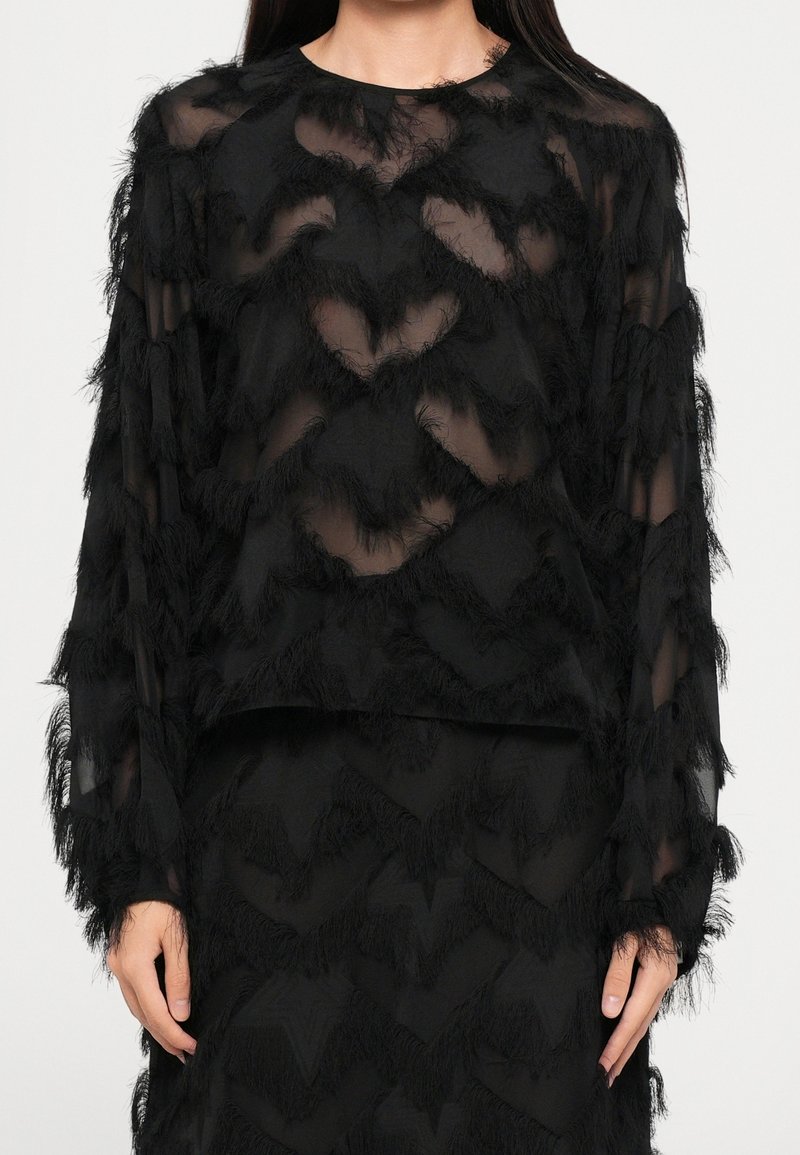 Femme portant un haut noir transparent à manches longues avec des franges en zigzag texturées et une jupe noire assortie.
