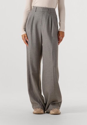 Grijze broek met wijde pijpen en plooien, gemaakt van gestructureerde stof; voorzien van riemlussen en voorzakken. Draagt beige sneakers.