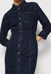 Denim knäppklänning i mörkblått med långa ärmar, elastisk midja, bröstficka och silverfärgade tryckknappar framtill.