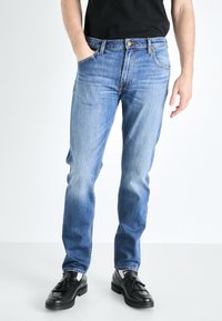 Light blue straight-leg jeans in denim fabric, featuring five pockets, a button closure, and subtle fading for texture. Worn with black shoes.