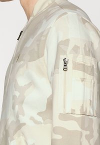 Giubbotto bomber mimetico beige con tasca con zip sulla manica e colletto a costine. Tessuto liscio con dettagli sottili di trama e motivo.