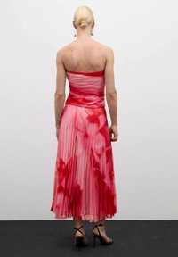 Strapless, geplooide midi-jurk in een rood en roze verloop met een tie-dye patroon. Heeft een getailleerde top en een vloeiende rok. Geko Combineerd met zwarte hakken.