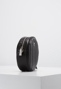 Borsa a tracolla nera in pelle trapuntata a forma ovale con cerniera argento e etichetta con logo, in posizione verticale su una piattaforma bianca contro uno sfondo chiaro.