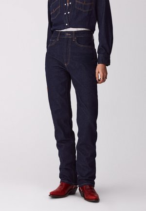 Person trägt dunkelblaue, hoch taillierte Jeans, passendes Jeanshemd und rote Lederstiefel und steht vor einem schlichten Hintergrund.