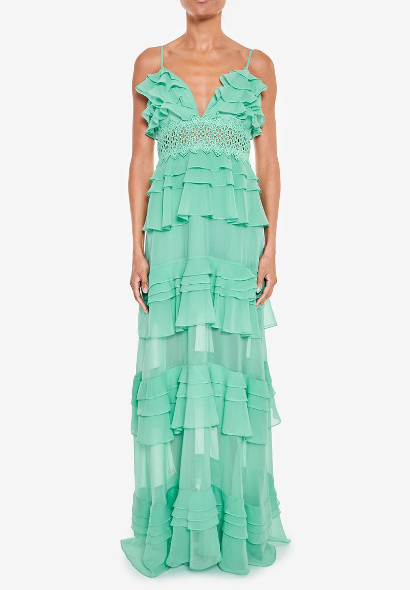 Femme portant une robe longue vert menthe avec des volants superposés, un empiècement en dentelle et des fines bretelles sur fond blanc.