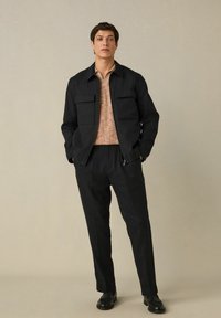 Men's black jacket with two front pockets, paired with a textured light brown shirt and black tailored pants. Worn with black footwear.