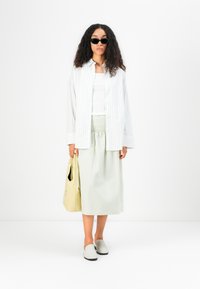 Stripet hvit skjorte med knapper, hvit tanktop, mintgrønn skjørt, lysegrå slip-on sko, svarte solbriller, og en gul tote bag.