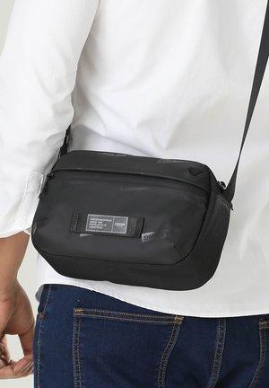 Sac bandoulière noir en matériau durable, présentant un design rectangulaire épuré, des compartiments zippés et une bandoulière.
