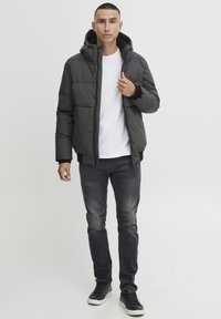 Puffer jacket in dark gray, quilted design, with a hood and elastic cuffs. Worn over a white T-shirt, paired with gray jeans and black sneakers.