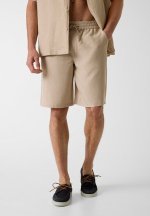 Homme portant un short beige à cordon, une chemise à manches courtes déboutonnée et des chaussures bateau noires avec des lacets marron, debout avec une main dans la poche.