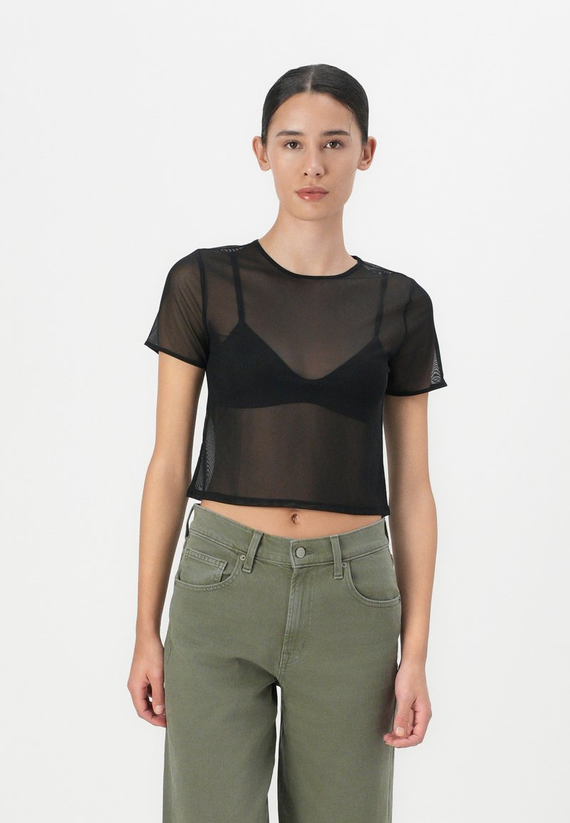 T-shirt cropped in rete nera con maniche corte, caratterizzato da un collo a giro e indossato sopra un bralette nero. Abbina a pantaloni larghi verde oliva.