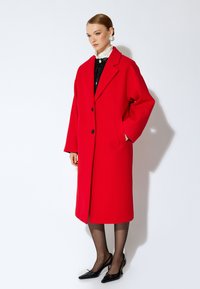Abrigo de lana rojo de gran tamaño con cierre de doble botonadura, amplios solapas acampanadas y bolsillos laterales. Estilizado sobre una blusa negra con lentejuelas y tacones negros.