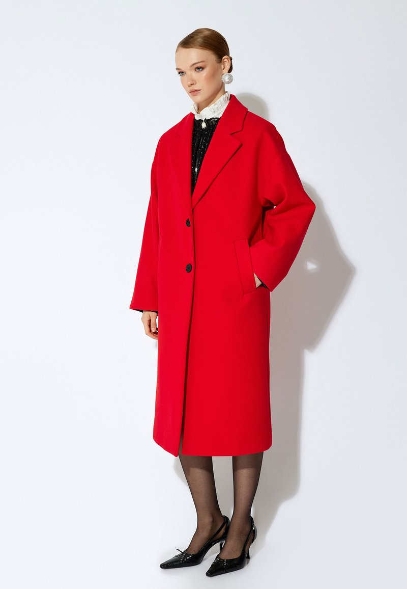 Abrigo de lana rojo de gran tamaño con cierre de doble botonadura, amplios solapas acampanadas y bolsillos laterales. Estilizado sobre una blusa negra con lentejuelas y tacones negros.