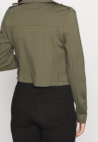 Giacca cropped verde oliva con maniche lunghe, colletto piatto e spalline. Tessuto liscio con un design strutturato. Pantaloni neri visibili sotto.