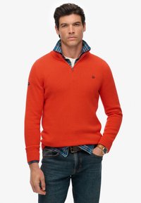 Pullover arancione lavorato a maglia con colletto a mezza zip, caratterizzato da un logo scuro sul petto, indossato sopra una camicia a quadri blu, abbinato a jeans blu.