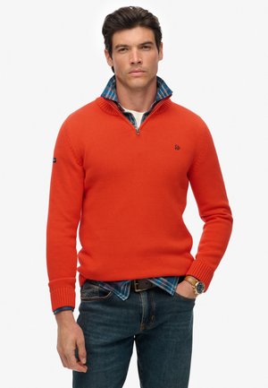 Suéter de punto naranja con un cuello de media cremallera, con un logotipo oscuro en el pecho, superpuesto a una camisa de cuadros azul, combinado con jeans azules.
