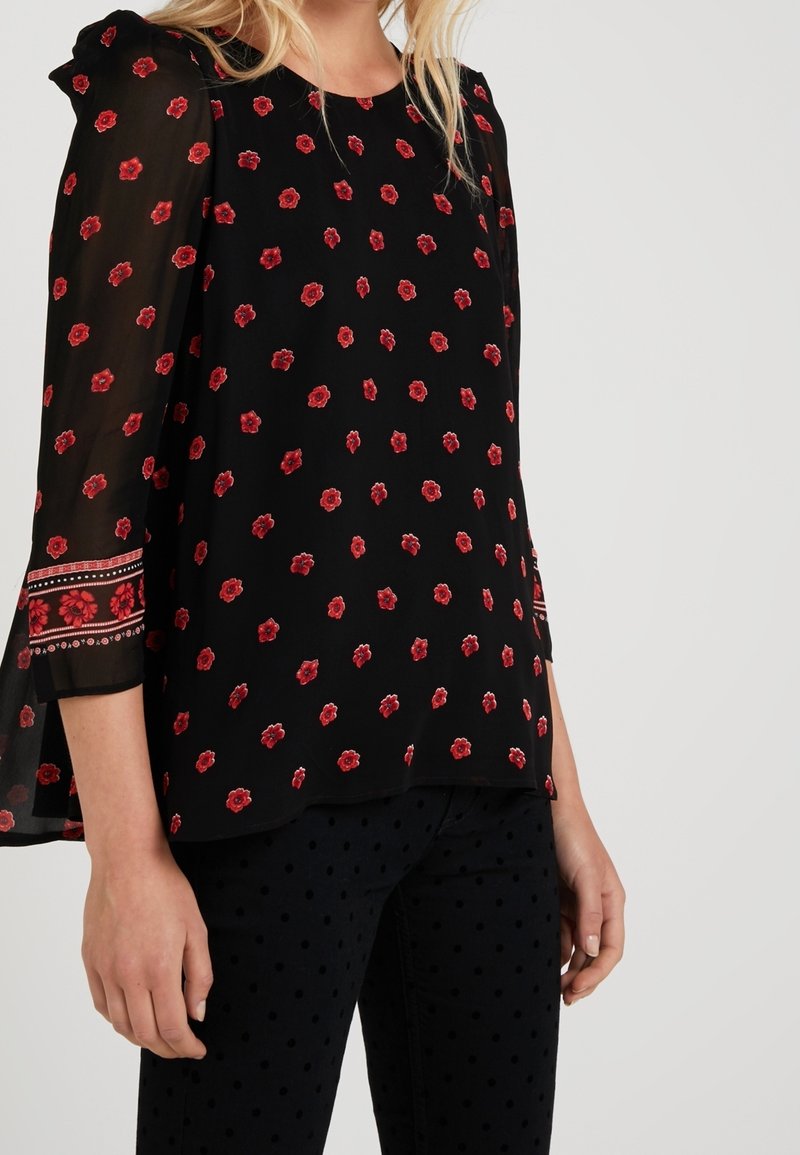 Blouse noire avec imprimé floral rouge, manches transparentes et design évasé. Présente une bordure décorative sur les bords des manches. Assortie avec un pantalon à pois.