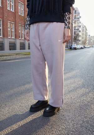 Persoană îmbrăcată cu pantaloni de trening roz deschis de la Nike, cizme negre masive și un pulover negru cu mâneci cu model, stând pe o stradă din oraș.