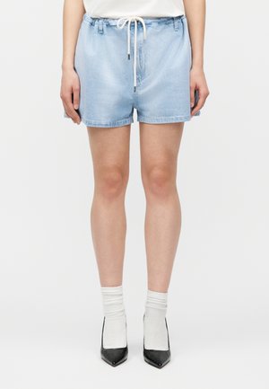 Άτομο που φορεί ανοιχτόχρωμα τζιν σορτς με λευκό κορδόνι, λευκές κάλτσες μέχρι τον αστράγαλο και μαύρα μυτερά ψηλοτάκουνα, στέκεται μπροστά σε λευκό φόντο.