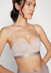 Lys beige bh med en kontureret sweetheart-udskæring, tynde stropper og en satinlignende bånd med logo. Glat stoftekstur.