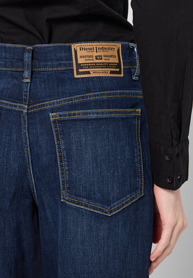 Jeans in denim blu scuro con un'etichetta in pelle marrone che presenta il logo "Diesel Industry". La tasca posteriore è contornata da cuciture gialle.