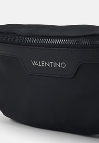 Musta nailonilaukku, jossa on sileä pinta, vetoketjulla varustettu päällinen ja hopeinen "VALENTINO" -logo etupaneelissa. Kompakti ja minimalistinen muotoilu.