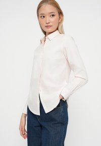 Camisa de botón de color rosa claro con mangas largas, cuello puntiagudo y dobladillo curvado. Combinada con jeans de mezclilla oscuros, mostrando un corte relajado.