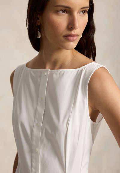Woman with long dark hair wearing sleeveless white buttoned top and shell-shaped earrings, looking to the side against a plain background.