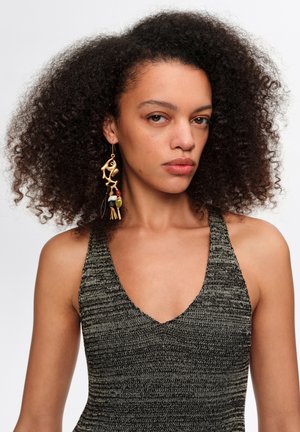 Mujer con cabello rizado y voluminoso que lleva un top gris de punto sin mangas y un gran pendiente colgante de oro y cuentas en su oreja izquierda.