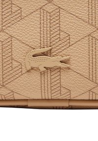 Μπεζ πορτοφόλι από εργασμένο δέρμα με γεωμετρικό ανάγλυφο μοτίβο και εμφανή ταμπά διακοσμητικό λογότυπο κροκοδείλου. Κομψός και δομημένος σχεδιασμός.