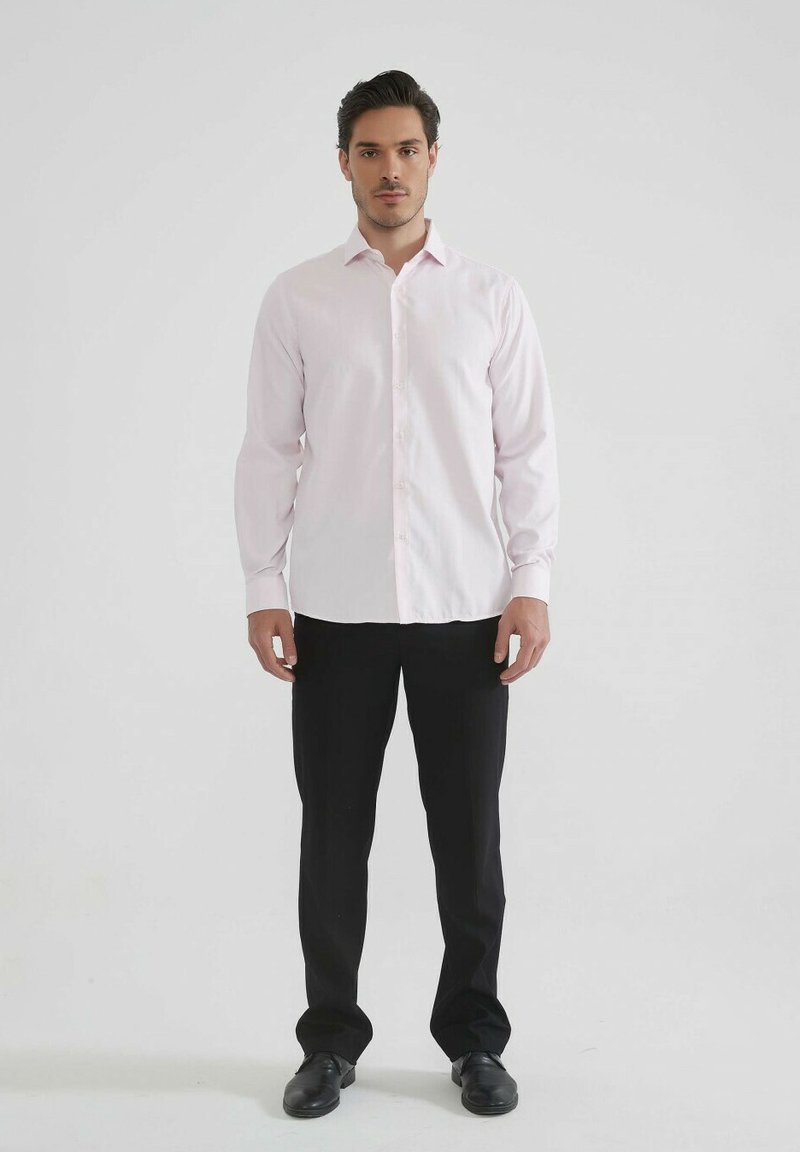 Chemise boutonnée à manches longues de couleur rose pâle avec un design ajusté. Associée à un pantalon noir et des chaussures noires, se tenant devant un fond uni.