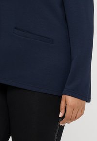 Haut à manches longues bleu marine au design simple, avec une poche frontale et une texture lisse, associé à des leggings noirs avec un accent brillant sur le côté.