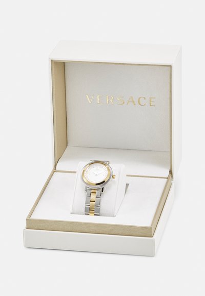 Auksinis ir sidabrinis laikrodis su apvaliu ciferblatu, baltu dialu, raštuotu ratlankiu ir dviejų spalvų metaline apyranke, gulintis baltoje dėžutėje su auksiniais akcentais.