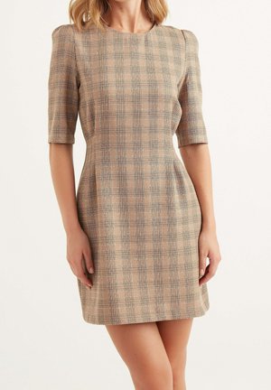 Kobieta ubrana w beżową, dopasowaną sukienkę w kratę z rękawami sięgającymi łokci i okrągłym dekoltem, stojąca na białym tle.