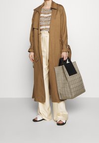 Brązowy długi płaszcz, paski w beżowym i brązowym kolorze na górze, kremowe szerokie spodnie, białe sandały, nosząc dużą torbę w kratę z czarnym uchwytem.