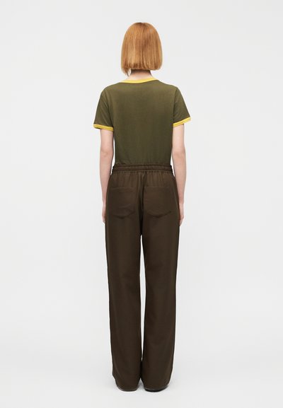 Σκούρο πράσινο μπλουζάκι με κίτρινη λεπτομέρεια, συνδυασμένο με χαλαρά καφέ παντελόνια που έχουν δύο πίσω τσέπες. Βλέπουμε την πλάτη, σε απλό φόντο.