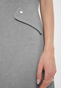 Grijze stoffen jurk met een klepzakje dat is vastgezet met een zilveren knop. Gladde textuur, getailleerde pasvorm, gedetailleerde stiksels, hand rustend nabij de zak.
