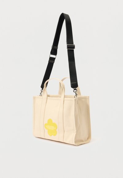 Bežová plátenná taška s dvojitými hornými rukoväťami a odnímateľným čiernym popruhom. Obsahuje logo žltej kvetiny a viditeľné švy. Obdĺžnikový tvar.