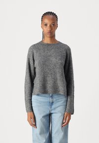 Pull en tricot gris avec un col rond, des manches longues et une coupe décontractée. Texture douce, style légèrement court, porté avec un jean bleu clair.
