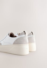 Zapatos sin cordones de ante y cuero en blanco y gris, con punta redonda y suela blanca gruesa, con costuras decorativas y tiradores en el talón.