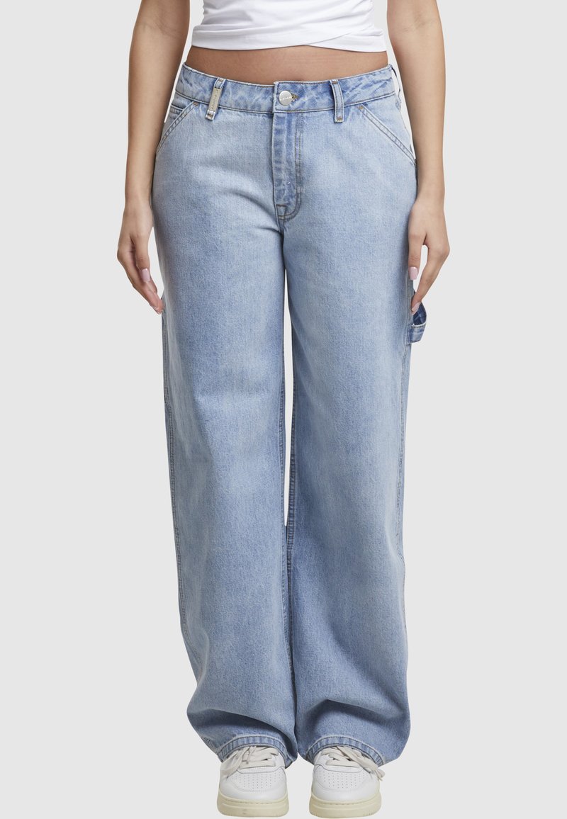 Personne portant un jean large bleu clair, des baskets blanches et un top blanc court, debout devant un fond clair et uni.