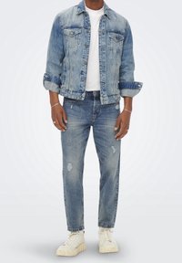 Vaaleansininen farkkutakki, jossa napillinen edusta ja kaksi rintataskua, yhdistettynä vaaleansinisiin kuluneisiin farkkuihin ja beigen värisiin paksupohjaisiin tennareihin.