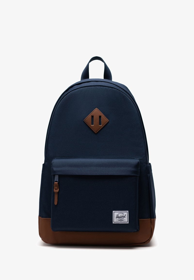 Tmavě modrý batoh zaobleného tvaru, s hnědými akcenty z umělé kůže, přední kapsou na zip a nášivkou v podobě diamantu na vrchu.
