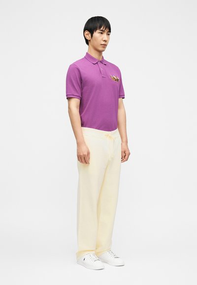 Άνδρας όρθιος, φορώντας μωβ πόλο μπλούζα με λογότυπο αλόγου, κρεμ παντελόνι με κορδόνι και λευκά αθλητικά παπούτσια μπροστά σε απλό λευκό φόντο.