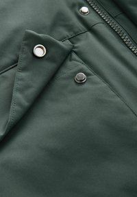 Jachetă de culoare verde închis, fabricată dintr-un material cu o textură netedă, cu capse argintii și un fermoar. Include un detaliu cu o clapetă triunghiulară pentru un plus de interes.