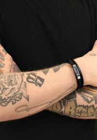 Bracciale in pelle intrecciata nera con piastra metallica con testo inciso, indossato su un braccio tatuato su uno sfondo di camicia nera.