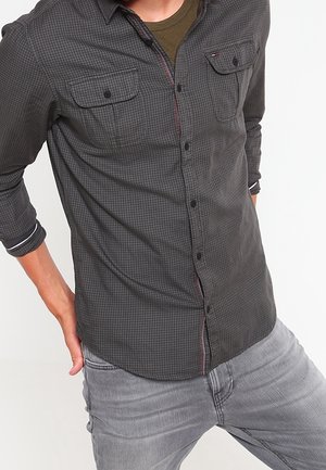 Mężczyzna w ciemnoszarej koszuli w kratę zapinanej na guziki, na oliwkowej zielonej koszulce i jasnoszarych dżinsach, stojący na białym tle.